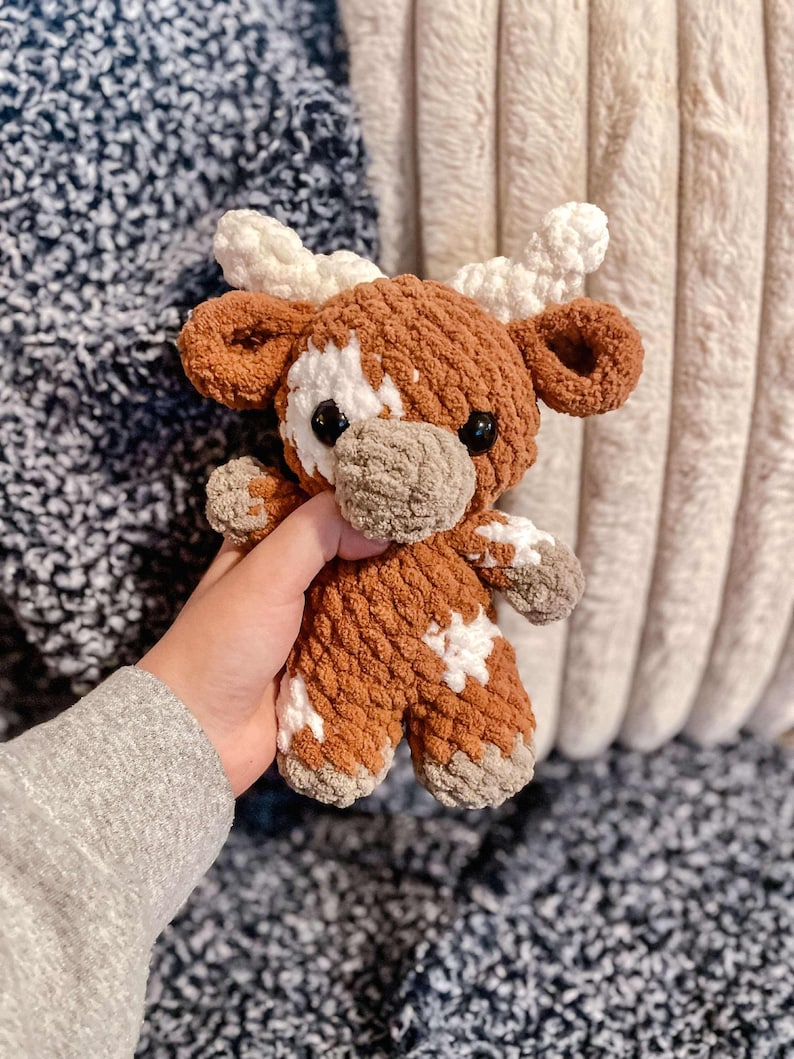 May include: A brown and white crocheted stuffed animal cow with a white star pattern. The cow has a brown nose and black eyes.