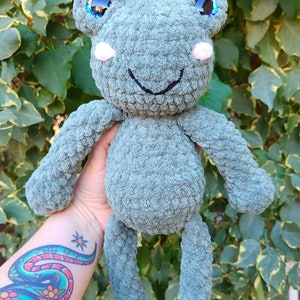 May include: A crocheted green frog plush toy with iridescent blue eyes and pink cheeks. The frog is smiling and has long legs.