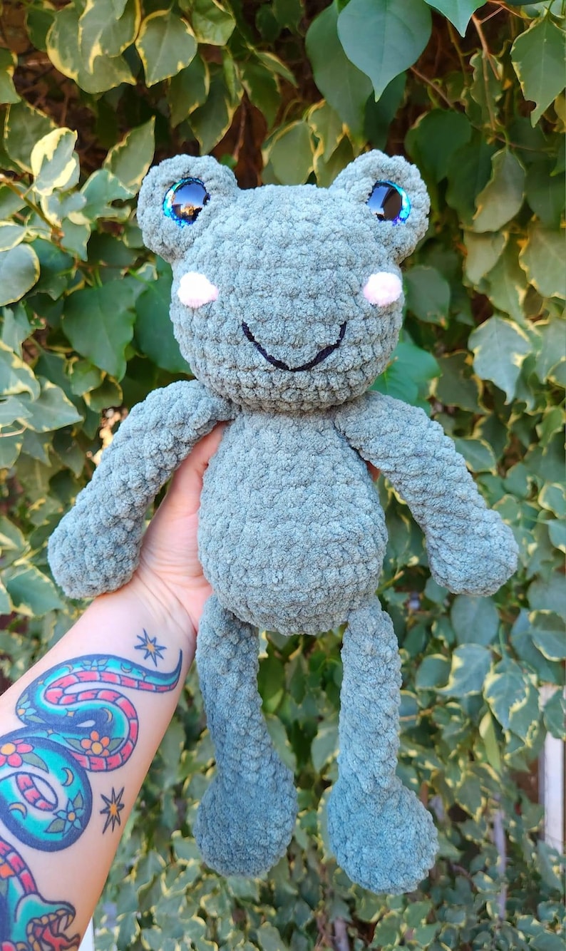 May include: A crocheted green frog plush toy with iridescent blue eyes and pink cheeks. The frog is smiling and has long legs.