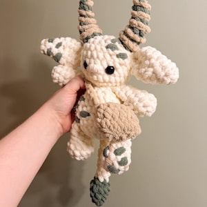 Peut inclure: Une peluche crochet&eacute;e &agrave; la main repr&eacute;sentant une cr&eacute;ature fantaisiste. Le jouet est principalement blanc cass&eacute; avec des d&eacute;tails verts et marron, notamment des taches, des cornes et un petit sac. La cr&eacute;ature a une expression amicale avec un &oelig;il en bouton noir.
