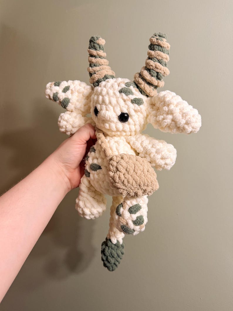 Peut inclure: Une peluche crochet&eacute;e &agrave; la main repr&eacute;sentant une cr&eacute;ature fantaisiste. Le jouet est principalement blanc cass&eacute; avec des d&eacute;tails verts et marron, notamment des taches, des cornes et un petit sac. La cr&eacute;ature a une expression amicale avec un &oelig;il en bouton noir.