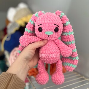 May include: A pink crocheted bunny with long, green and pink striped ears. The bunny has black eyes and a light pink nose.