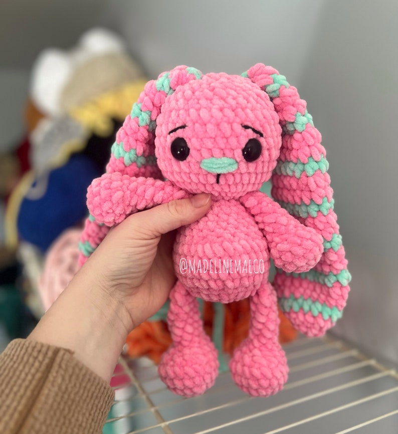 May include: A pink crocheted bunny with long, green and pink striped ears. The bunny has black eyes and a light pink nose.