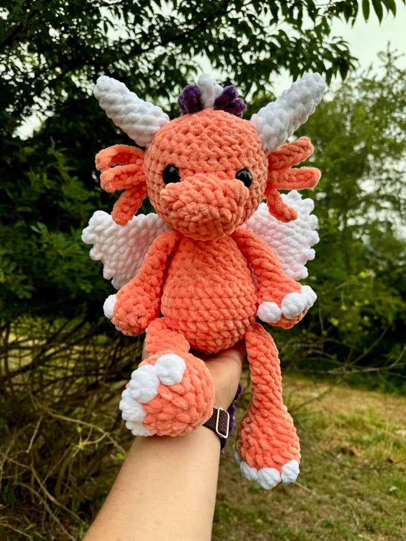 May include: A crocheted plush dragon toy with orange fur, white wings, and white horns. The dragon has black eyes and a purple flower on its head.
