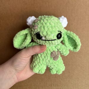 Könnte beinhalten: Ein kleines grünes Häkeltier aus Plüsch mit weißen Hörnern und schwarzen Augen. Das Spielzeug hat einen braunen Fleck an der Seite und ein gesticktes Lächeln.