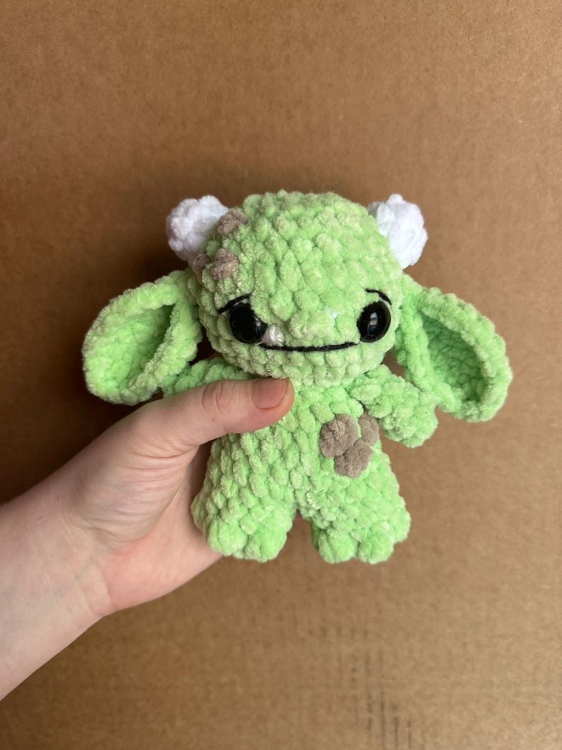 May include: A small green crocheted plush toy with white horns and black eyes. The toy has a brown patch on its side and a stitched-on smile.