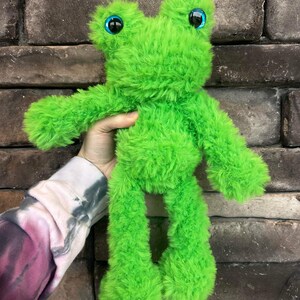 May include: A bright green plush frog toy with large blue eyes. The frog is standing on its hind legs and has a fluffy texture.