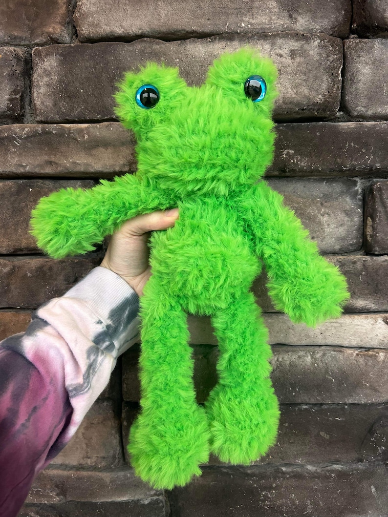 May include: A bright green plush frog toy with large blue eyes. The frog is standing on its hind legs and has a fluffy texture.