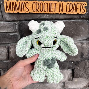 May include: Green and white crocheted plush toy with a friendly face and a heart-shaped design on its chest.  "Mama's Crochet N Crafts" is written on a sign behind the toy.