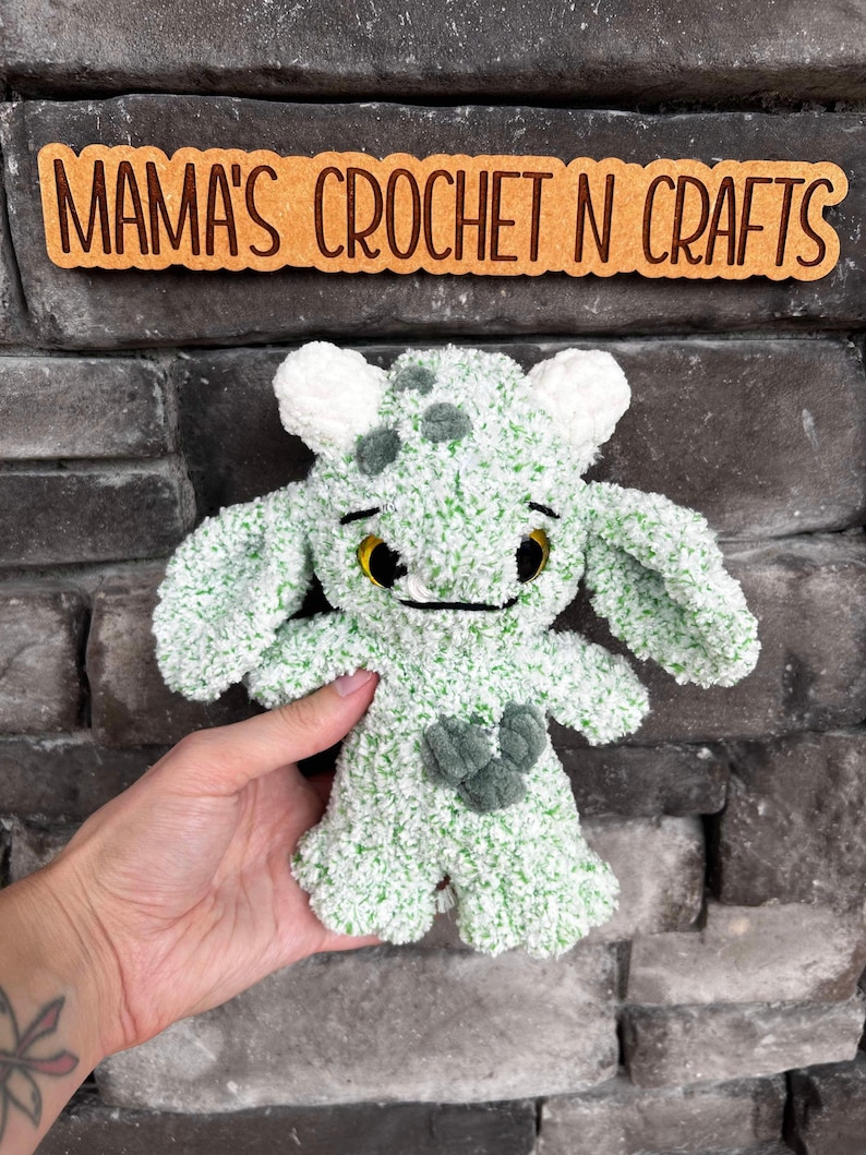 May include: Green and white crocheted plush toy with a friendly face and a heart-shaped design on its chest.  "Mama's Crochet N Crafts" is written on a sign behind the toy.