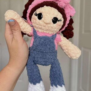 May include: A crocheted doll with brown hair, a pink bow, and a blue denim overall dress. The doll has a light brown face with black eyes and a pink mouth.
