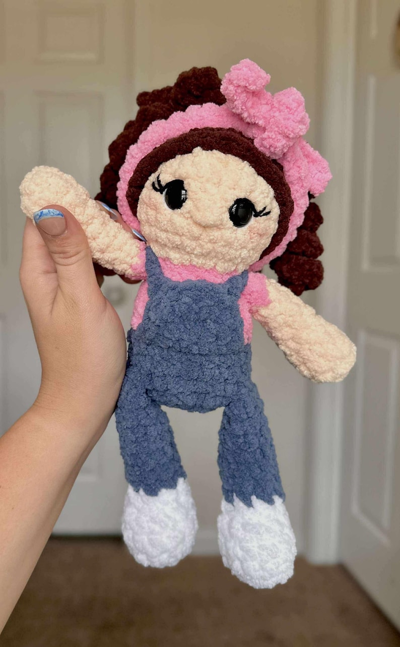 May include: A crocheted doll with brown hair, a pink bow, and a blue denim overall dress. The doll has a light brown face with black eyes and a pink mouth.