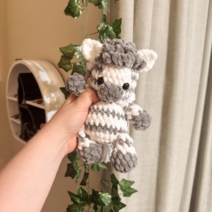 Peut inclure: Une peluche zèbre crochetée à la main, blanche et grise. Le zèbre a un corps rayé, une crinière grise et de petites oreilles arrondies. La peluche est douce et câline, idéale pour un jouet d'enfant ou une décoration de chambre d'enfant.
