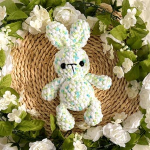 May include: A handmade crochet bunny in white with blue and green flecks, set on a woven basket. The bunny has black eyes and a black nose. The background features a wreath of white flowers and green leaves.