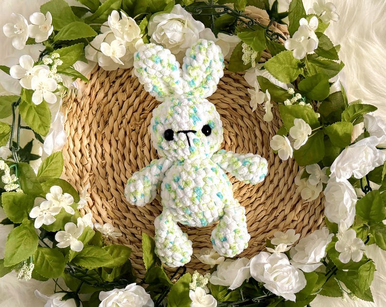 May include: A handmade crochet bunny in white with blue and green flecks, set on a woven basket. The bunny has black eyes and a black nose. The background features a wreath of white flowers and green leaves.