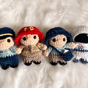 May include: Four crocheted plush toys, each representing a different profession. The first is a police officer with a blue uniform and a yellow badge. The second is a firefighter with a red helmet and a yellow and brown uniform. The third is a mail carrier with a blue uniform and a blue bag. The fourth is an astronaut with a white and blue spacesuit.