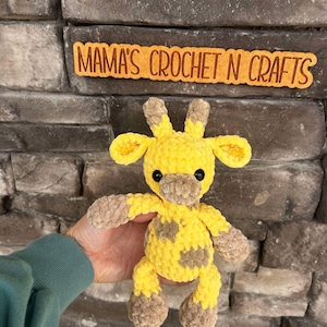 May include: A handmade yellow and brown crochet giraffe with black safety eyes. The giraffe has brown spots and is being held in front of a stone wall. The sign above the giraffe reads "MAMA'S CROCHET N CRAFTS".