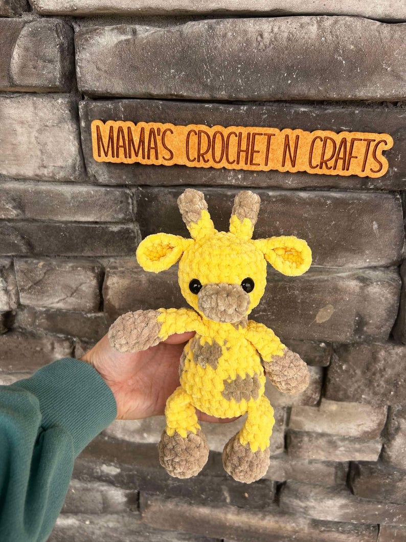 May include: A handmade yellow and brown crochet giraffe with black safety eyes. The giraffe has brown spots and is being held in front of a stone wall. The sign above the giraffe reads "MAMA'S CROCHET N CRAFTS".
