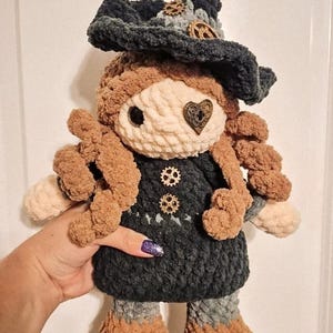 May include: A handmade crochet doll with a whimsical witch design. The doll features a dark blue hat adorned with gears, a cream-colored face, and brown curly hair. The dress is dark blue with gear-shaped buttons. The doll is approximately 10 inches tall.