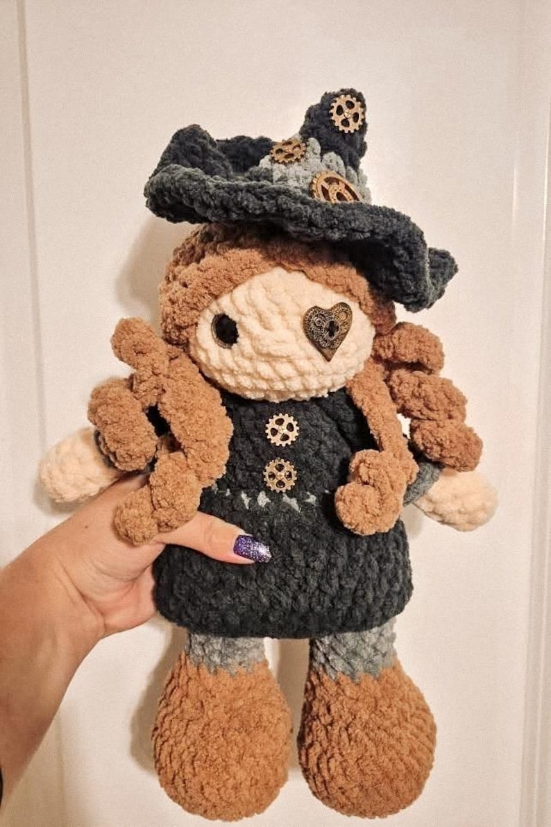 May include: A handmade crochet doll with a whimsical witch design. The doll features a dark blue hat adorned with gears, a cream-colored face, and brown curly hair. The dress is dark blue with gear-shaped buttons. The doll is approximately 10 inches tall.