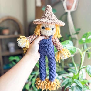 Könnte beinhalten: Handgemachte Häkelpuppe eines Vogelscheuche mit einem beigefarbenen Hut und strohähnlichen Haaren und Akzenten. Die Puppe trägt eine blaue Latzhose und hat ein aufgenähtes Gesicht. Die Puppe ist etwa 25 cm groß.