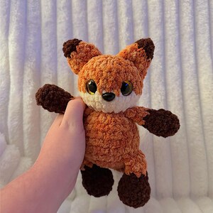 May include: A crocheted fox toy in orange and brown. The fox has large eyes, a white muzzle, and dark brown details on its ears, paws, and feet. The plush toy is soft and cuddly, suitable for children or as a decorative item. The fox is being held.