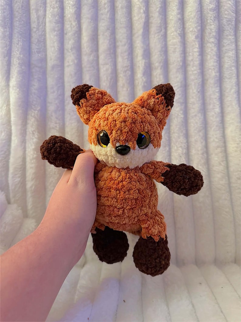 May include: A crocheted fox toy in orange and brown. The fox has large eyes, a white muzzle, and dark brown details on its ears, paws, and feet. The plush toy is soft and cuddly, suitable for children or as a decorative item. The fox is being held.