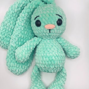 May include: A crocheted green bunny rabbit plush toy with black eyes and a pink nose. The bunny has long floppy ears and is sitting on a white background.
