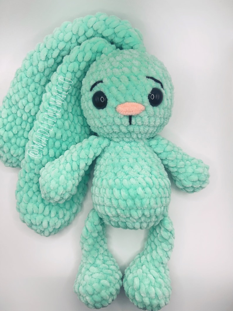 May include: A crocheted green bunny rabbit plush toy with black eyes and a pink nose. The bunny has long floppy ears and is sitting on a white background.