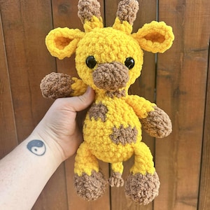 May include: A handmade yellow and brown crochet giraffe plush toy with black safety eyes. The giraffe has brown spots, ears, and hooves. The plush is held in a person's hand against a wooden background.