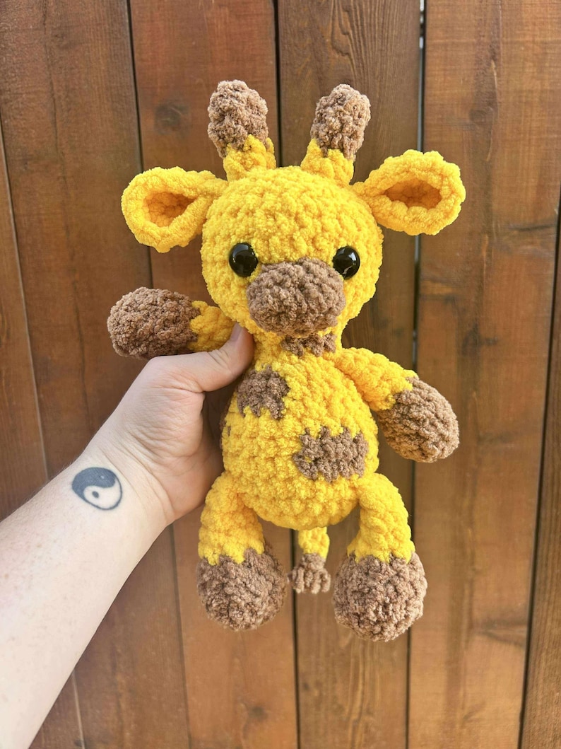 May include: A handmade yellow and brown crochet giraffe plush toy with black safety eyes. The giraffe has brown spots, ears, and hooves. The plush is held in a person's hand against a wooden background.