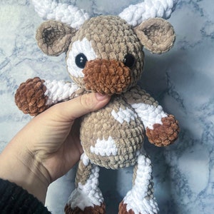 May include: A handmade crochet cow plushie in shades of brown, white, and tan. The plush toy has large black eyes, a brown muzzle, and white horns. The cow's body is a soft, textured material, and it is held by a hand.