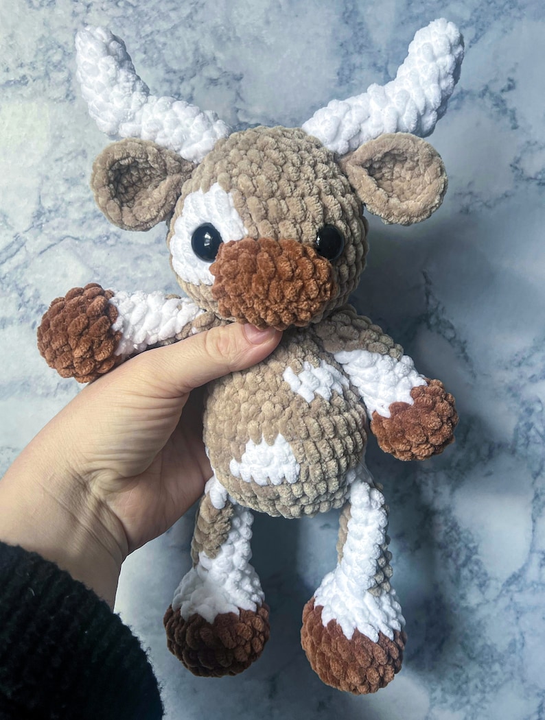 May include: A handmade crochet cow plushie in shades of brown, white, and tan. The plush toy has large black eyes, a brown muzzle, and white horns. The cow's body is a soft, textured material, and it is held by a hand.