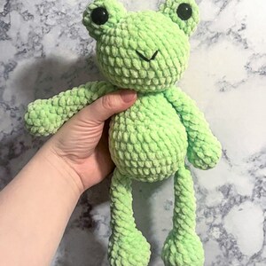 May include: A green crocheted frog plush toy with black eyes. The frog has a small smile and long legs.