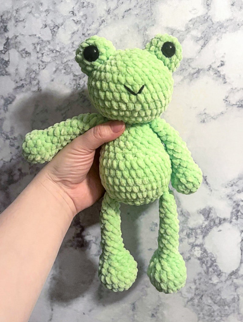 May include: A green crocheted frog plush toy with black eyes. The frog has a small smile and long legs.