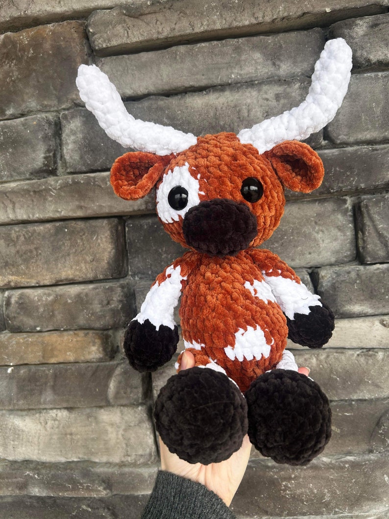 May include: A handmade crochet longhorn plush toy. The plush features a brown and white body with black hooves and a black snout. The horns are white. The toy is soft and cuddly, perfect for a child's room or as a decorative item.