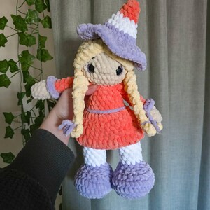 May include: A handmade crochet doll with a witch theme. The doll has long blonde hair, a red dress, and a purple witch hat. The doll's hat is striped with white and orange. The doll's feet and arms are purple and white.