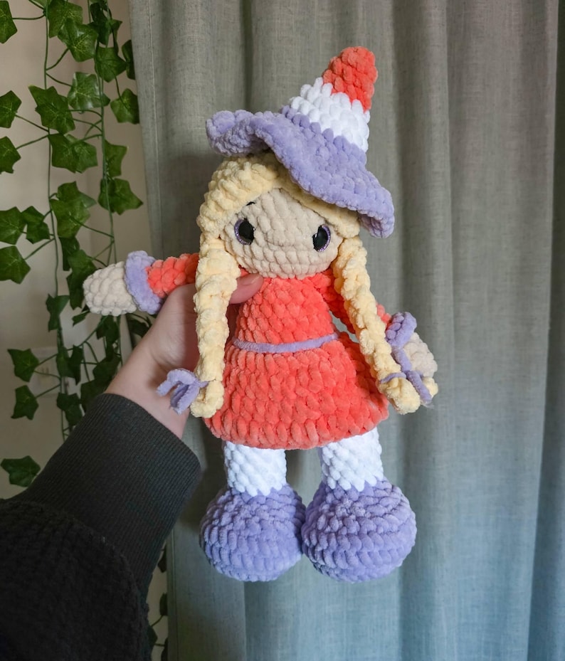 May include: A handmade crochet doll with a witch theme. The doll has long blonde hair, a red dress, and a purple witch hat. The doll's hat is striped with white and orange. The doll's feet and arms are purple and white.