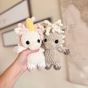 May include: Two crocheted stuffed unicorn toys. One is white with a pink and yellow mane and horn. The other is beige with a white mane and horn.