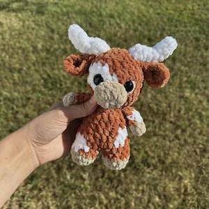 May include: A crocheted brown and white cow plush toy with white horns. The toy has black eyes and a beige nose.