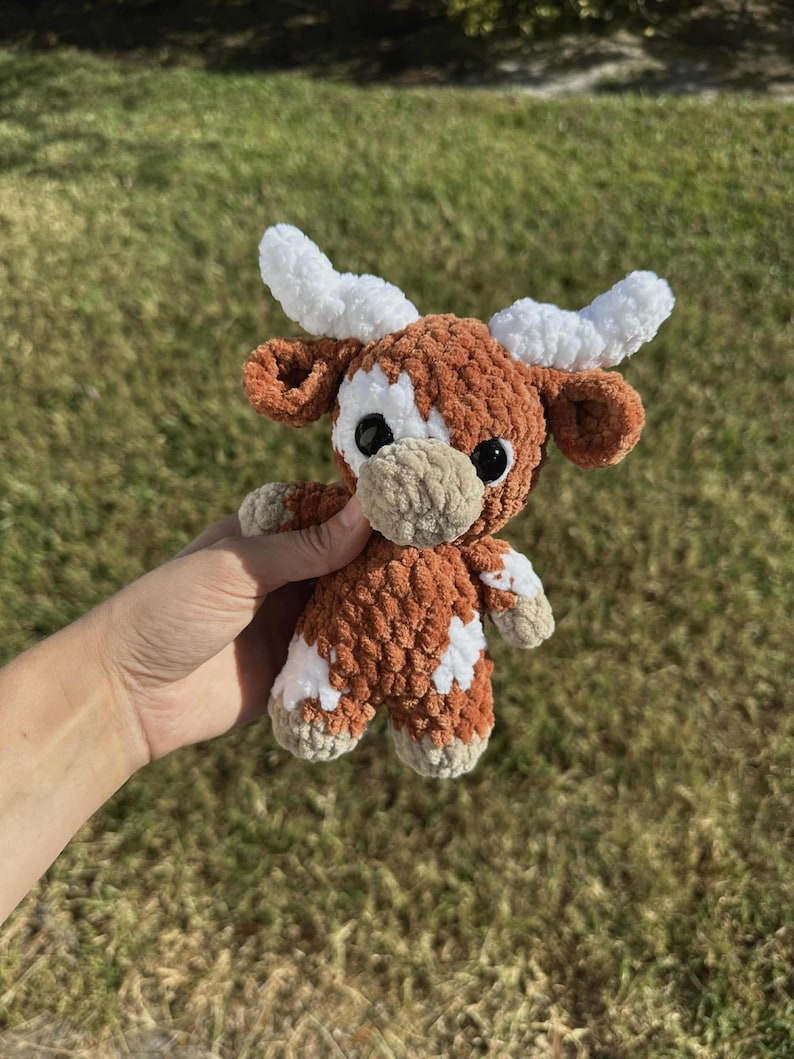 May include: A crocheted brown and white cow plush toy with white horns. The toy has black eyes and a beige nose.