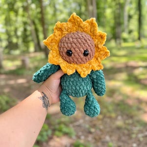 K&ouml;nnte beinhalten: Ein geh&auml;keltes Pl&uuml;sch-Sonnenblumenspielzeug mit braunem Gesicht und gr&uuml;nem K&ouml;rper. Das Spielzeug wird von einer Hand gehalten.