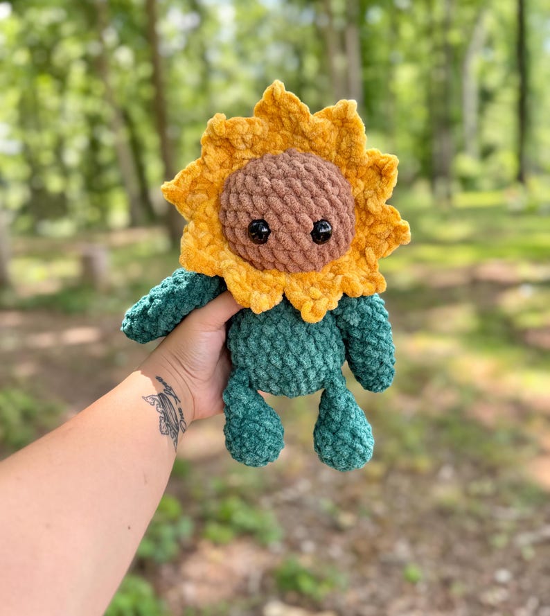 K&ouml;nnte beinhalten: Ein geh&auml;keltes Pl&uuml;sch-Sonnenblumenspielzeug mit braunem Gesicht und gr&uuml;nem K&ouml;rper. Das Spielzeug wird von einer Hand gehalten.