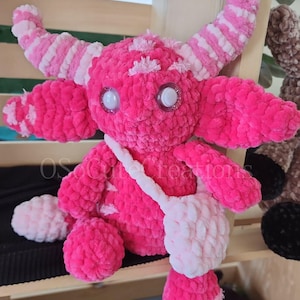 Peut inclure: Une peluche monstre rose faite &agrave; la main, avec de grands yeux scintillants, des cornes et un petit sac. La peluche est faite de fil doux et textur&eacute; et pr&eacute;sente une combinaison de rose uni et de d&eacute;tails roses et blancs ray&eacute;s. Le design g&eacute;n&eacute;ral est fantaisiste et mignon.