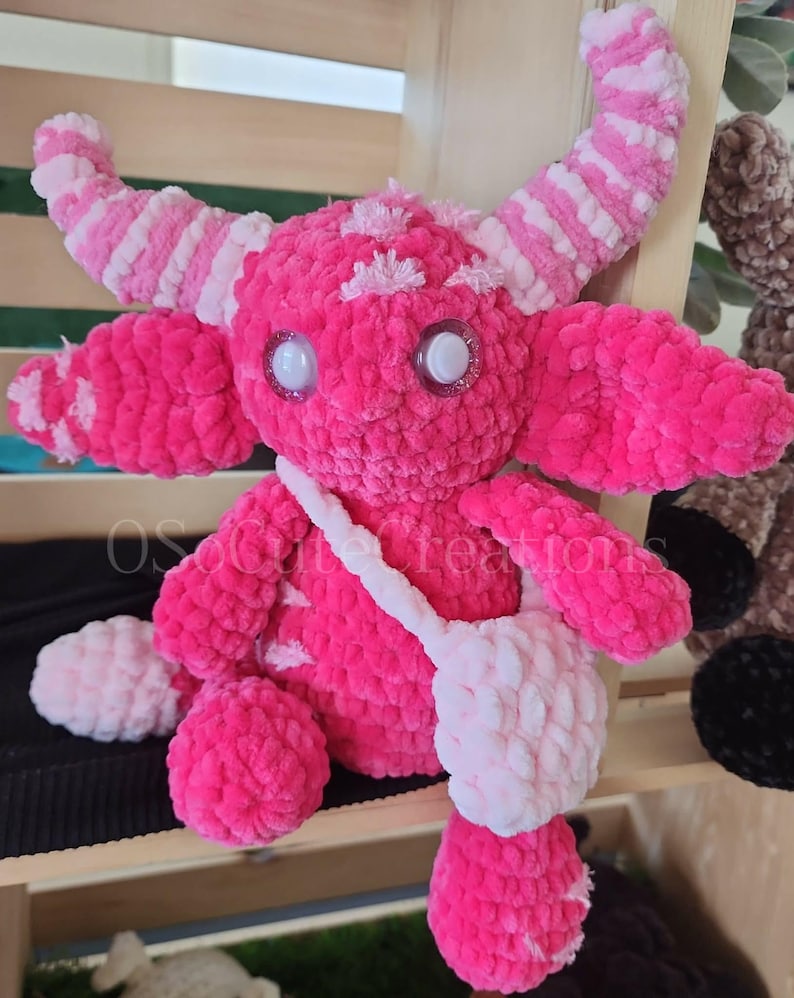 Peut inclure: Une peluche monstre rose faite &agrave; la main, avec de grands yeux scintillants, des cornes et un petit sac. La peluche est faite de fil doux et textur&eacute; et pr&eacute;sente une combinaison de rose uni et de d&eacute;tails roses et blancs ray&eacute;s. Le design g&eacute;n&eacute;ral est fantaisiste et mignon.