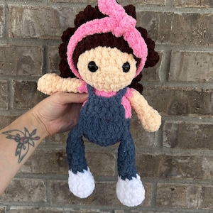 May include: A crocheted doll with brown hair, a pink headband, a blue overall, and white shoes.