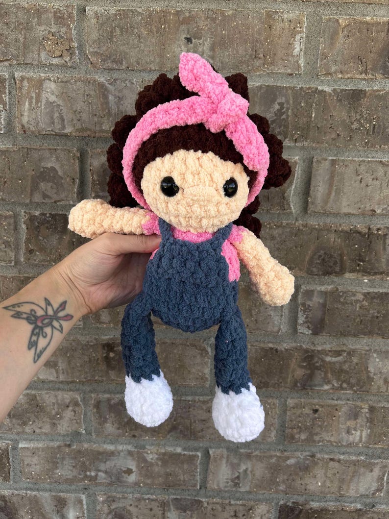 May include: A crocheted doll with brown hair, a pink headband, a blue overall, and white shoes.