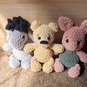 May include: Three handmade crochet plush toys featuring a grey donkey with a black mane, a yellow bear with a black nose, and a pink pig with a green shirt. Each stuffed animal is crafted from soft, textured yarn with black plastic safety eyes.