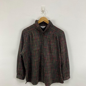 Könnte beinhalten: Langärmliges Hemd mit einem Karomuster in Grün-, Rot- und Brauntönen. Das Hemd hat einen hohen Kragen und hängt an einem Holzbügel. Der Stoff scheint ein strukturiertes Material zu sein.