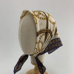 May include: A white and blue patterned headscarf with a gold chain design. The scarf is tied on a white mannequin head.
